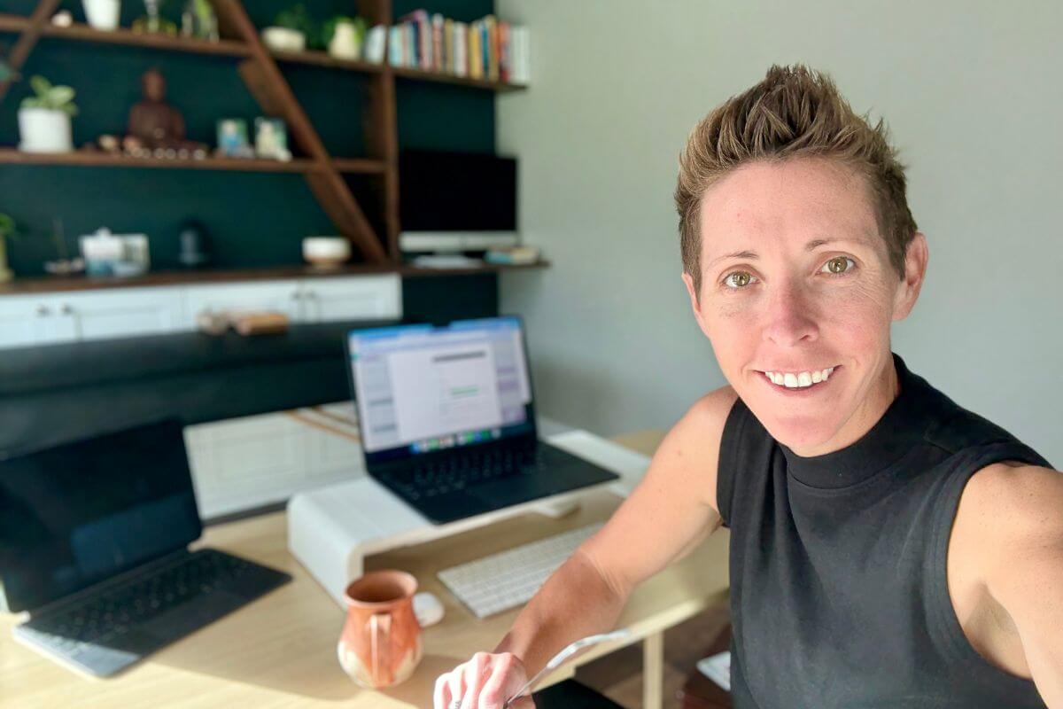 Kenzie Bauer smiling at her desk in a bright, modern home office with her laptop and coffee mug — representing the Feed the Purpose section of Feed the Birds First.