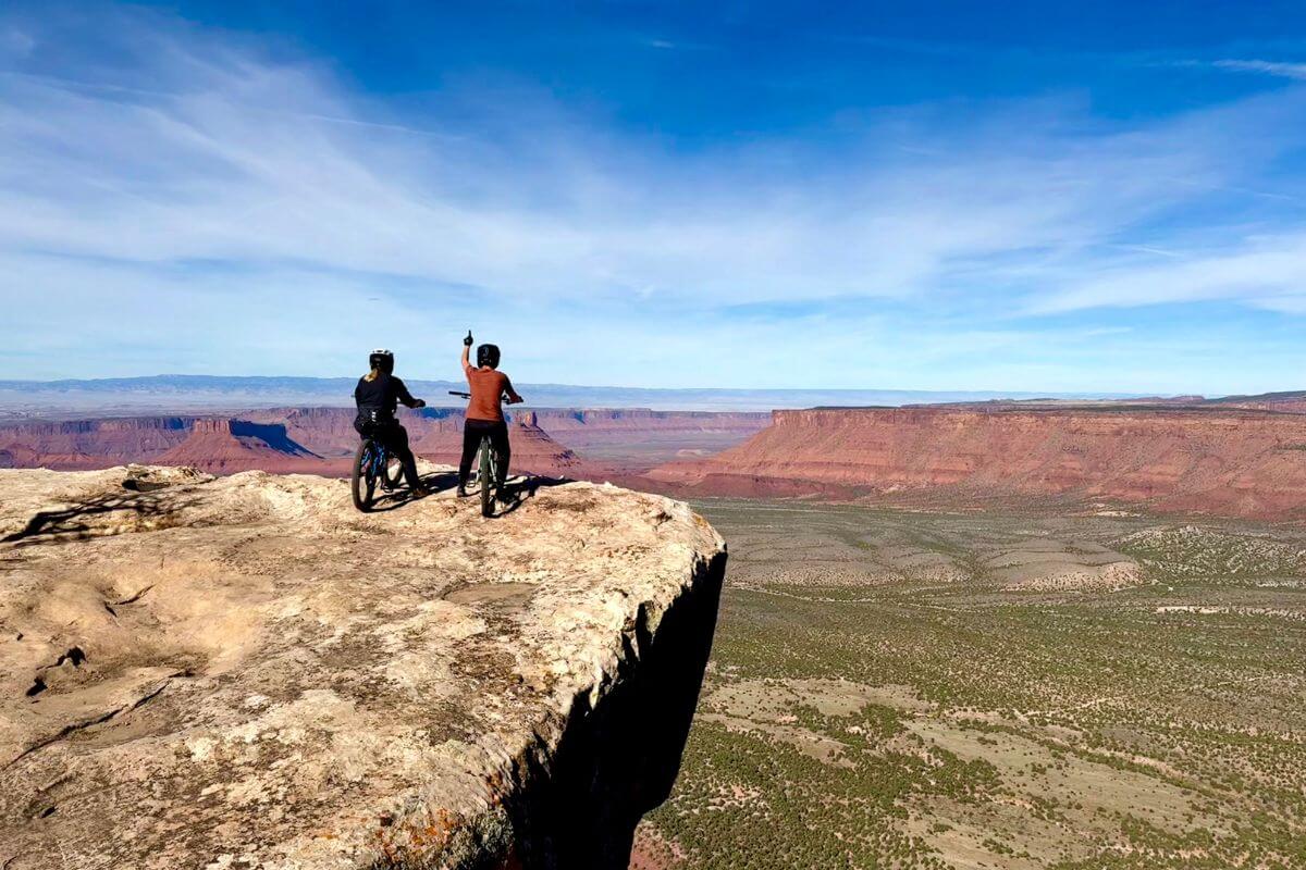 Two mountain bikers stand on the edge of a red rock cliff overlooking the vast desert canyons of Moab, Utah, under a bright blue sky.