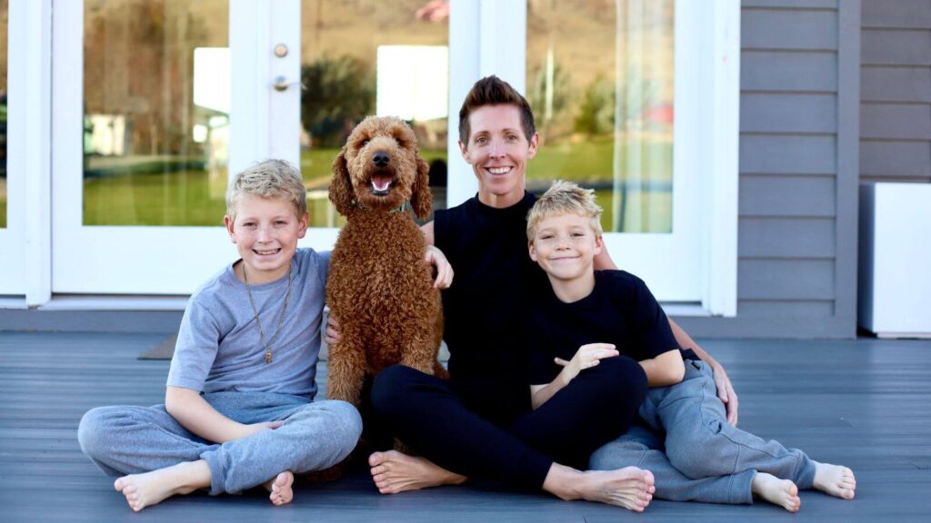 A smiling woman sitting on a porch with two young boys and a fluffy brown dog, all barefoot and relaxed together in front of large windows